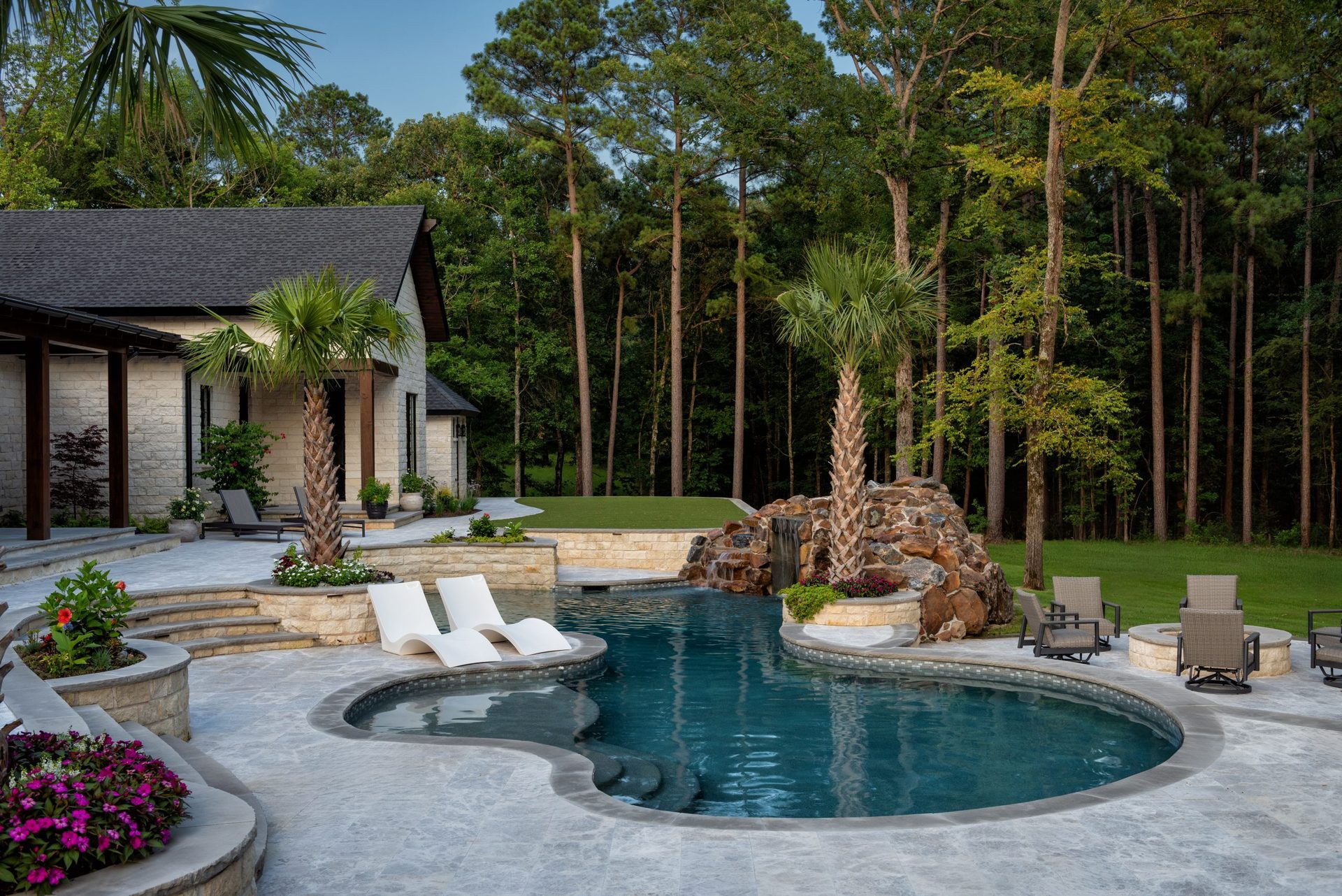 A freeform backyard pool with white lounge chairs on a travertine deck